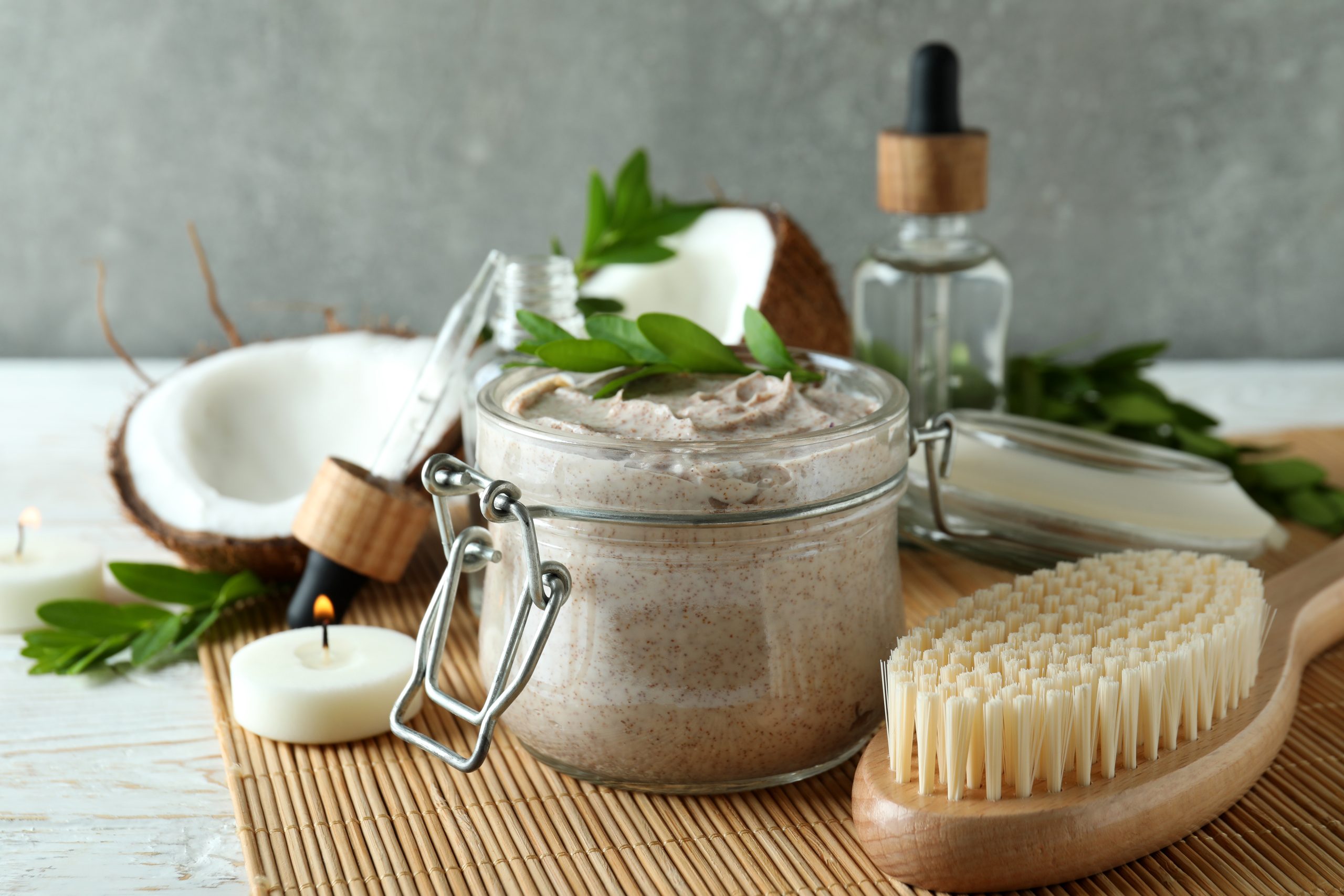 A bottle of skin care scrub in the centre and surround by essential oil, brush, candle, leaves and coconut